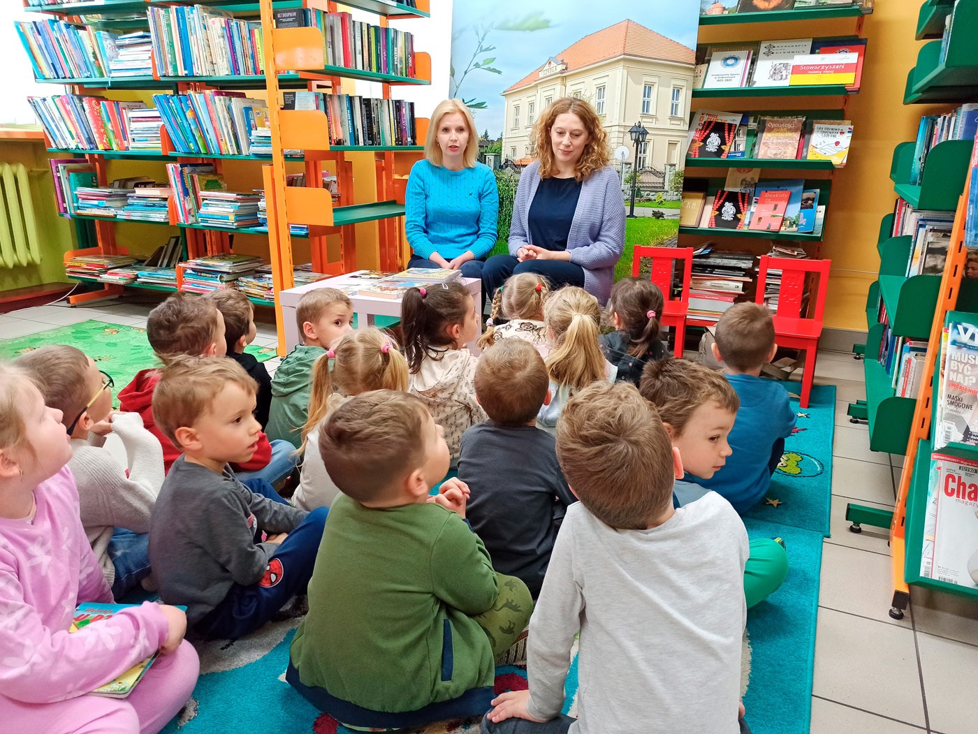 Na zdjęciu wdać grupę dzieci siedząca naprzeciw dwóch pań bibliotekarek, które opowiadają o książkach