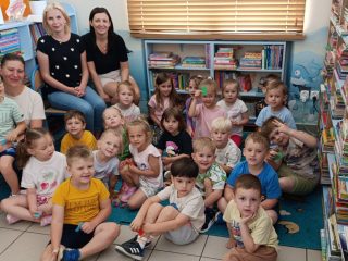 Prowadząca i wychowawczyni siedzą z tyłu na krzesłach, przed nimi dzieci siedzą na dywanie, twarze skierowane w stronę aparatu - zdjęcie grupowe.