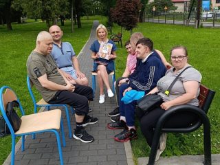 Osoby siedzą w parku na ławce i stołkach. Za nimi są drzewa i zielona trawa.